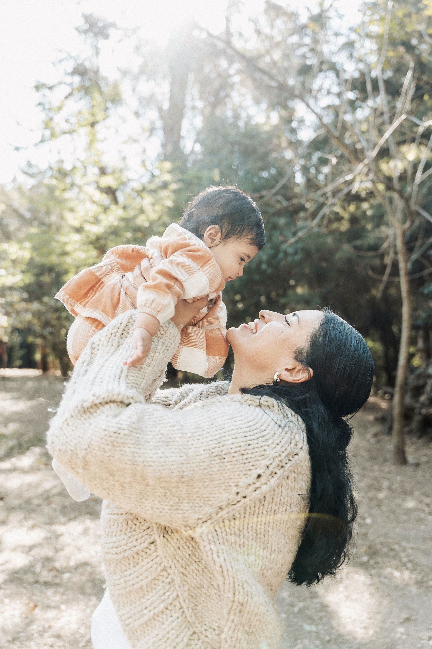 A parent in a cream knit sweater lifts their baby wearing a peach-colored outfit in a sunlit outdoor setting.