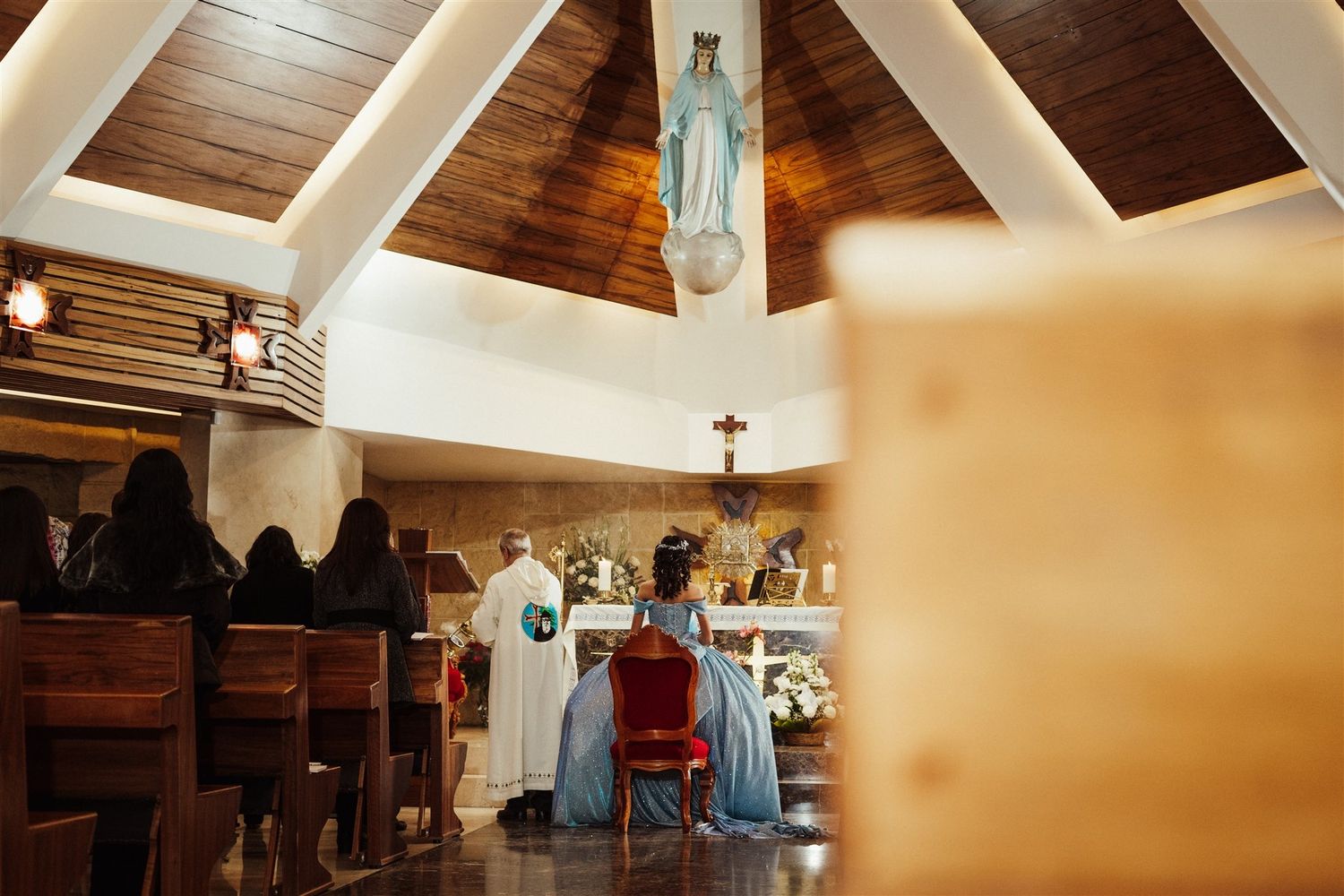 A Catholic church service takes place in a wooden-beamed chapel with a statue of the Virgin Mary mounted above.