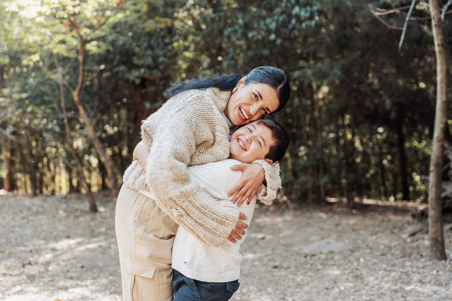 Two people in beige sweaters share a joyful embrace while laughing together in a sunlit forest setting.