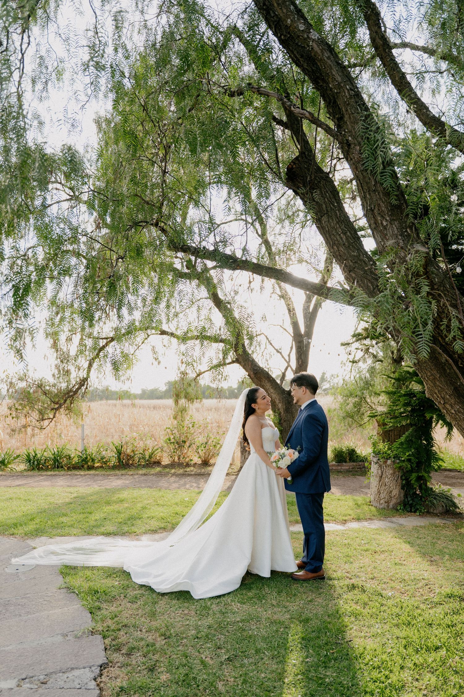A couple shares an intimate moment under a weeping willow tree during their outdoor wedding photoshoot.