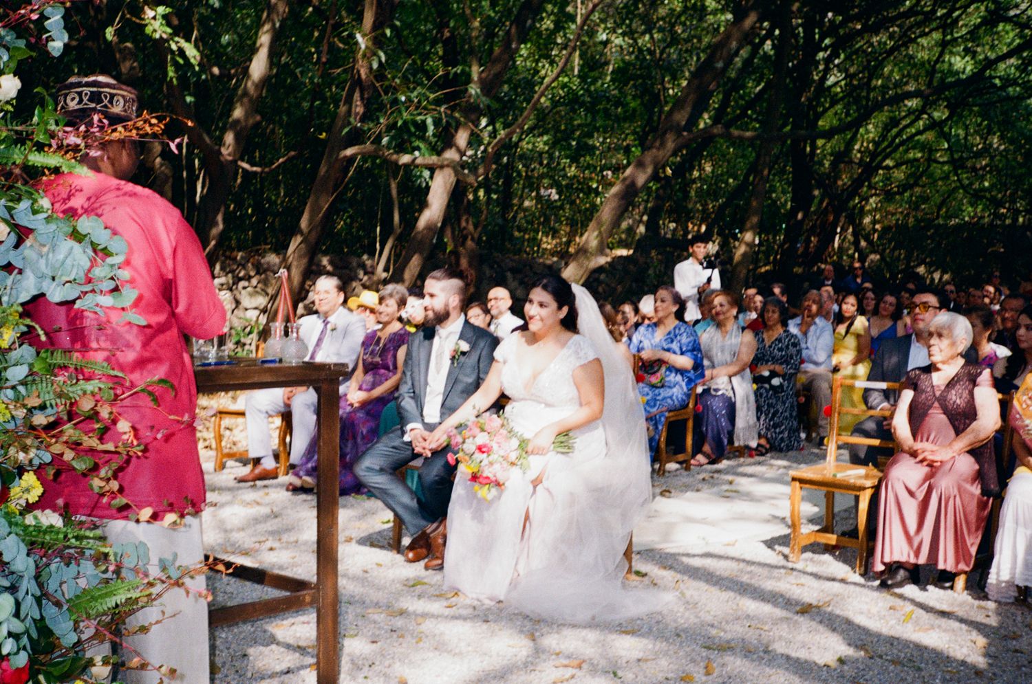 An outdoor wedding ceremony takes place in a lush garden setting with wooden benches and natural decor.