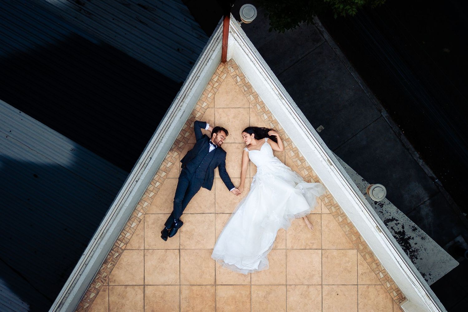 A bride and groom lying on tiled floor captured from above in triangular architectural framing.