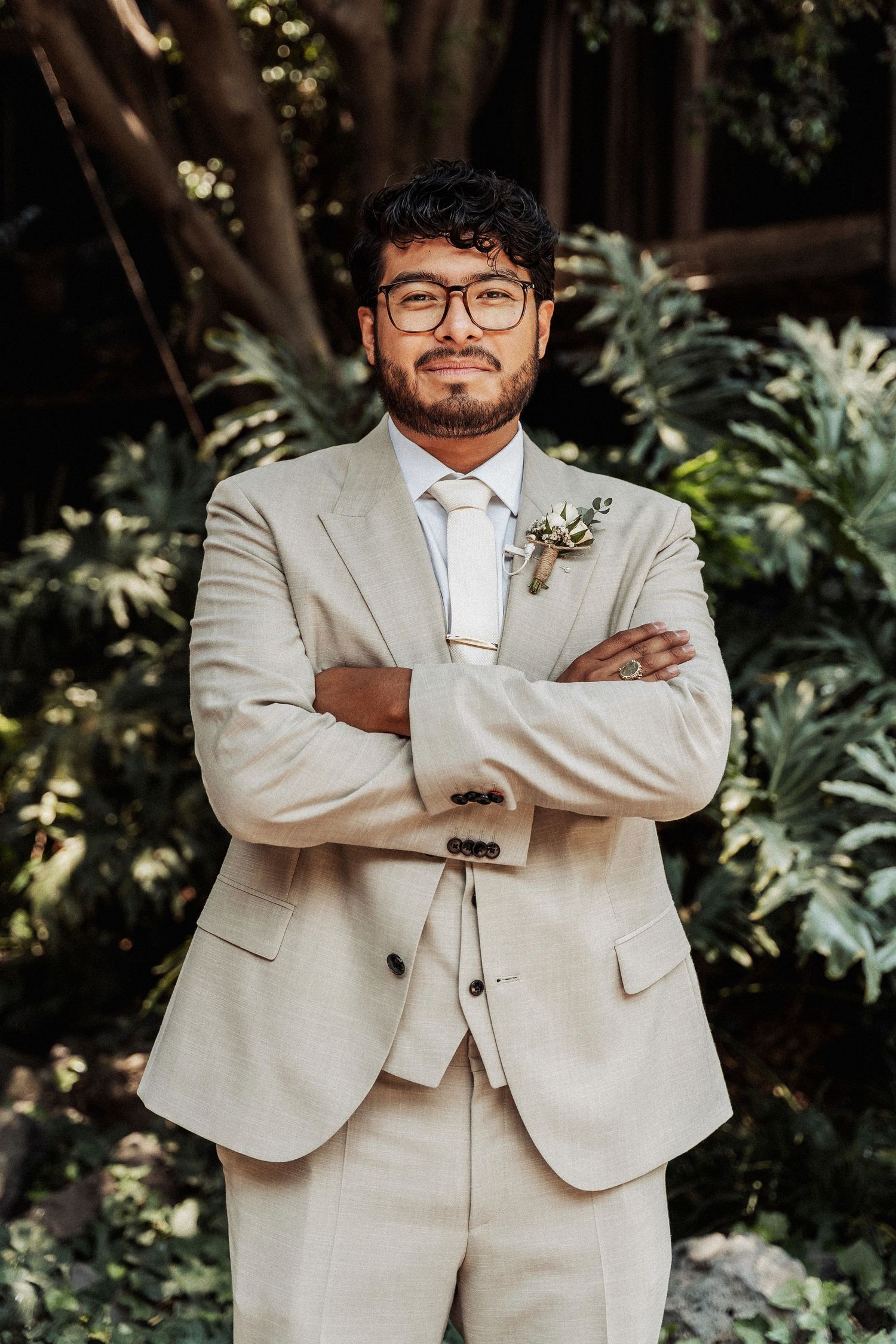 A person wearing a light beige suit and glasses stands in a garden setting with arms crossed.