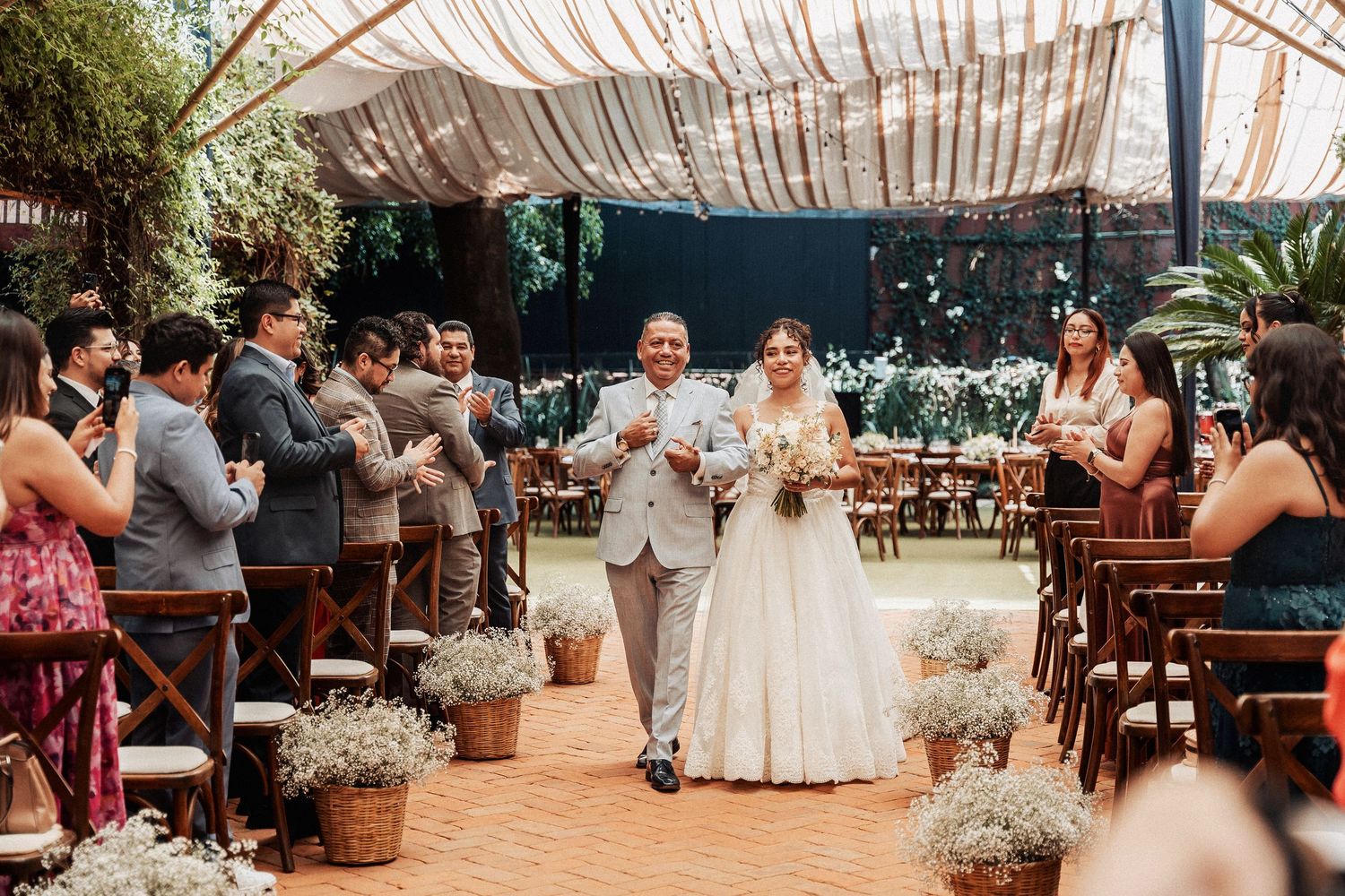 Happy couple walks down aisle at outdoor wedding ceremony under draped canopy with rustic floral arrangements.