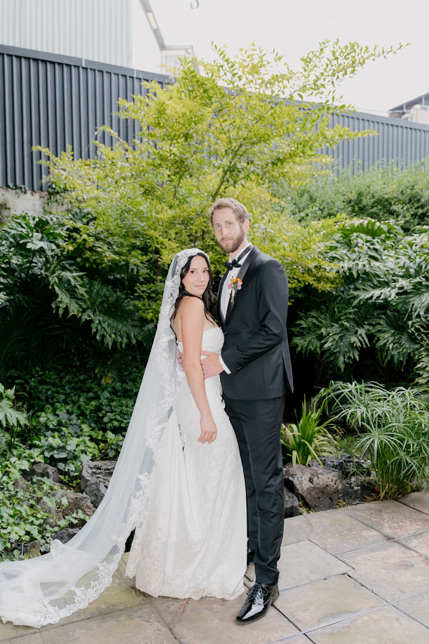 A romantic wedding couple poses in front of lush greenery in an outdoor garden setting.