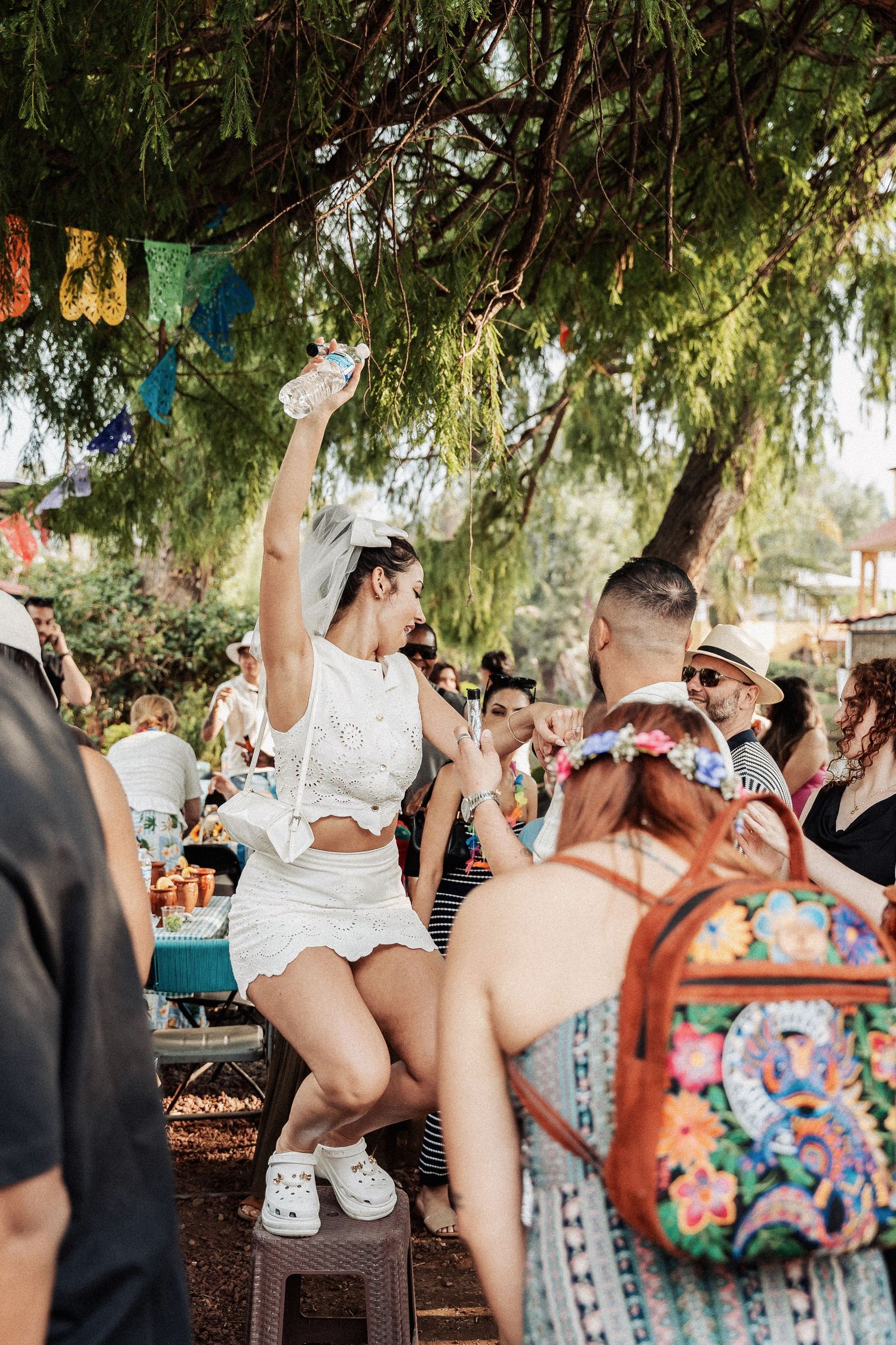 Outdoor festival scene with dancers in white outfits celebrating under trees with colorful decorations and banners.