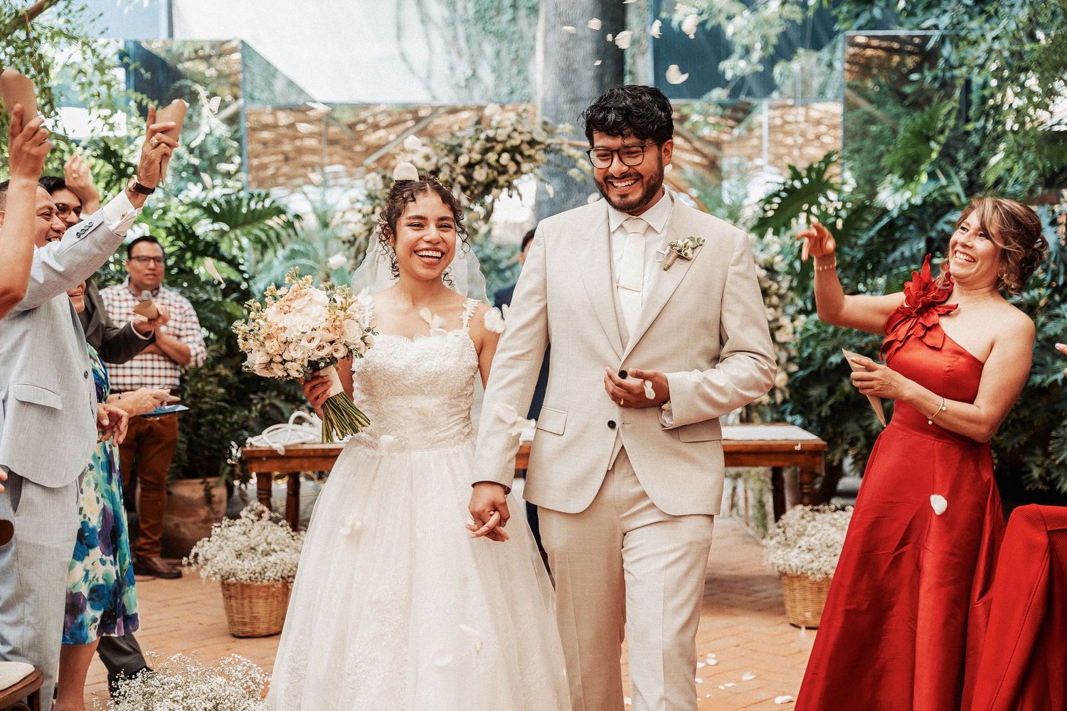 Wedding party in red bridesmaid dresses and tan suits celebrate joyfully in an outdoor garden setting.