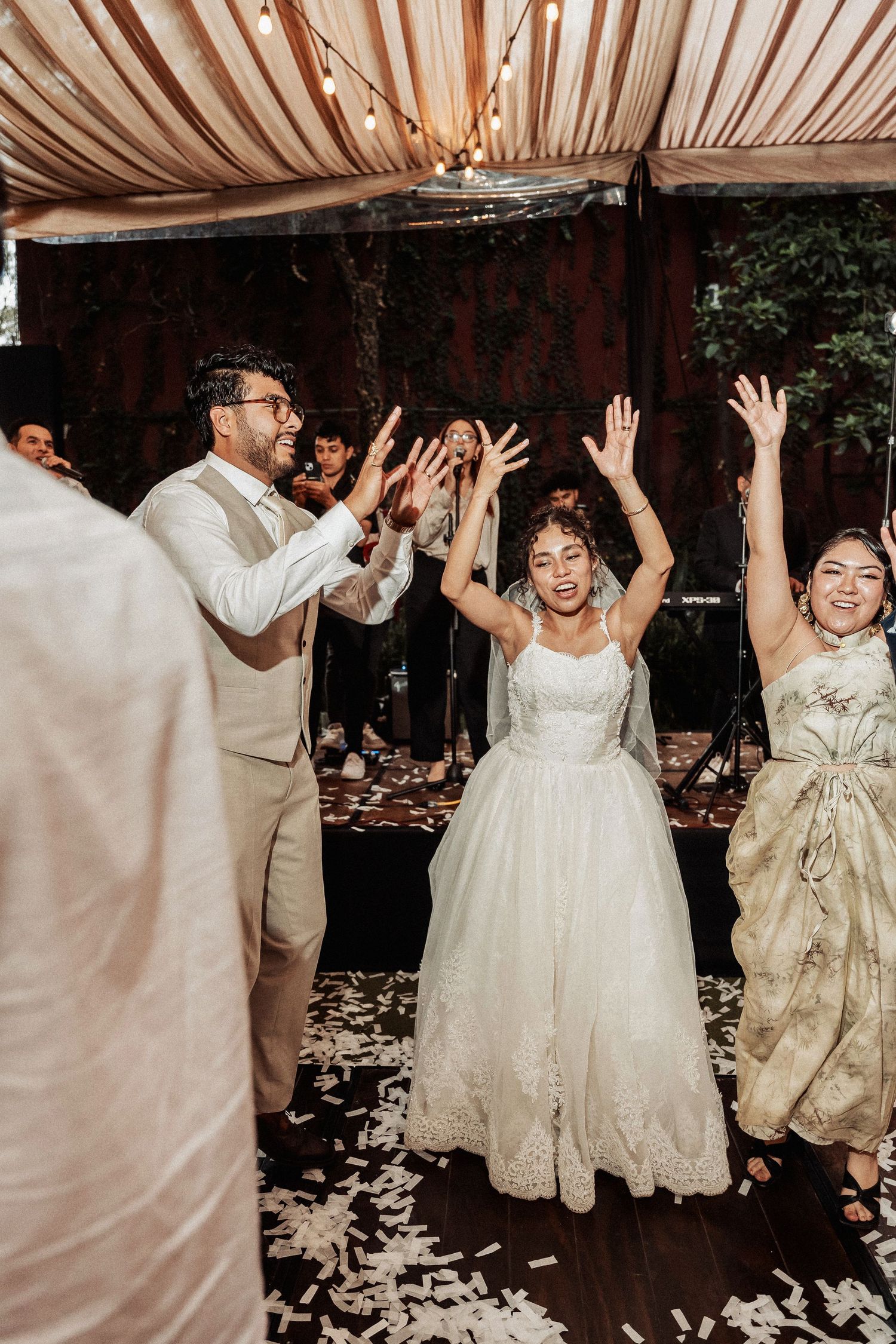 Wedding celebration with joyful dancing and raised hands on a dance floor decorated with flower petals under draped fabric.