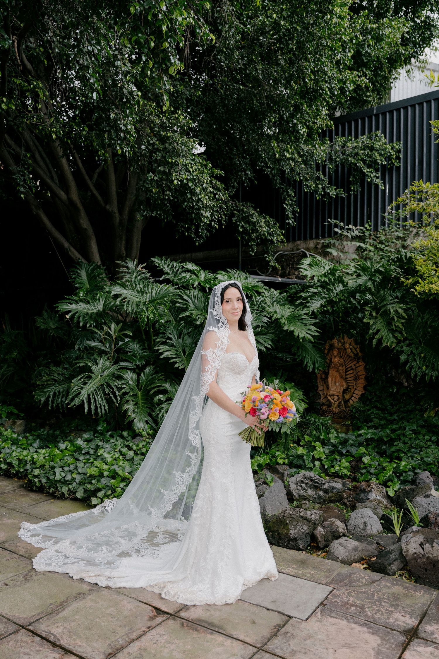 A bride in a lace wedding gown and long veil holds a vibrant bouquet while standing in a lush garden setting.