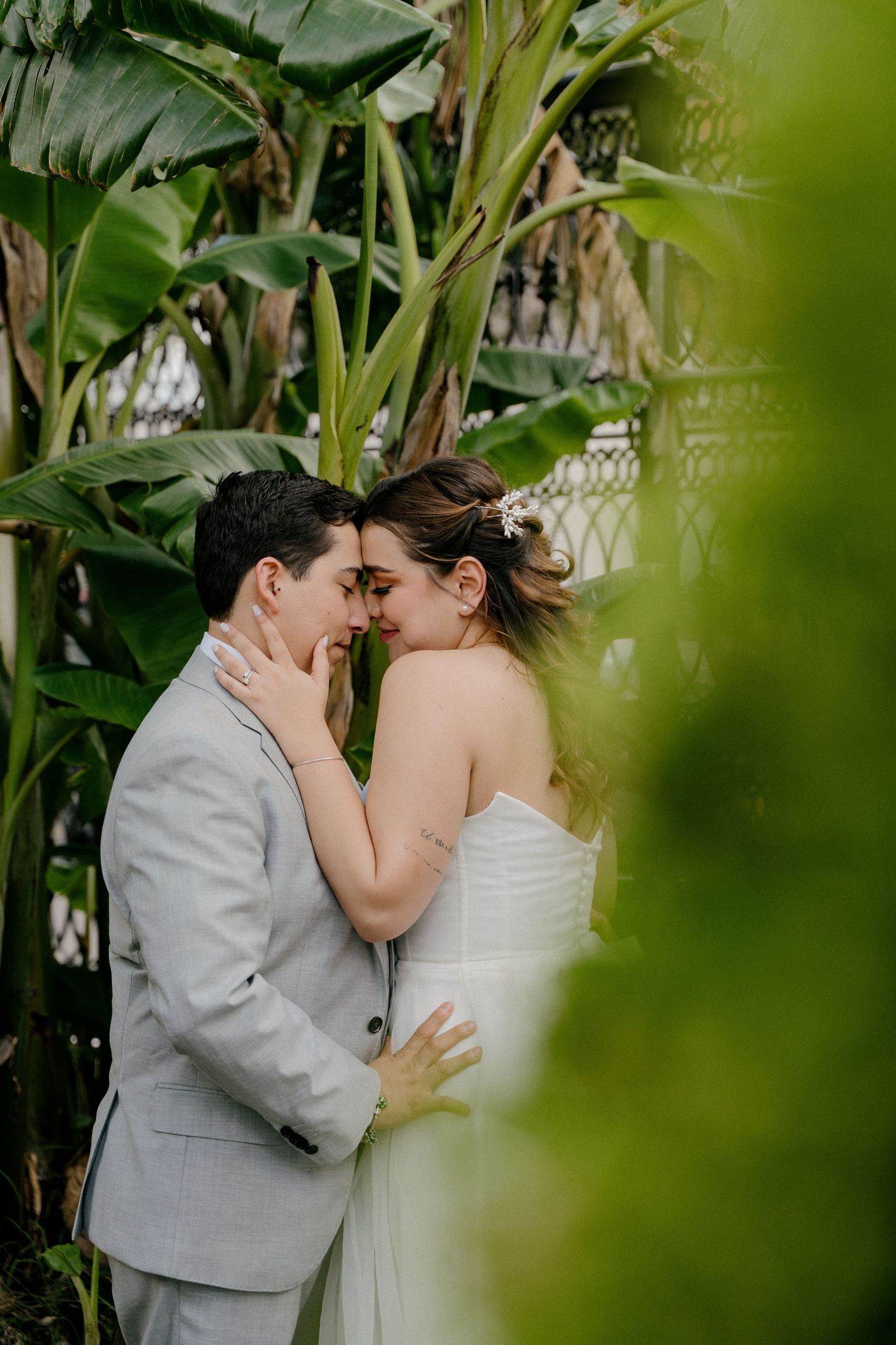 A romantic wedding moment captured through green foliage as couple shares intimate kiss in garden setting.