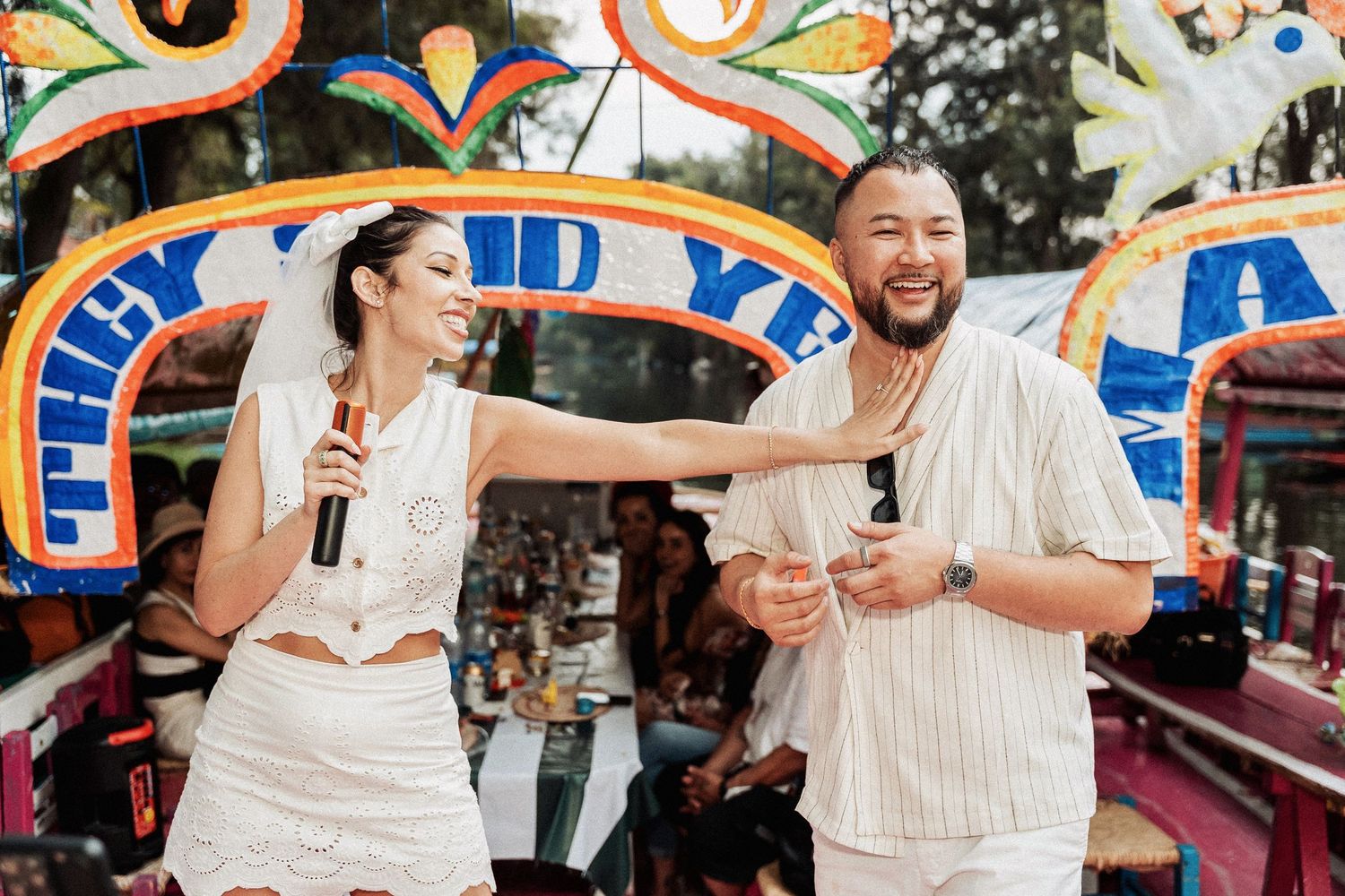 Two people in white outfits share a joyful laugh while holding microphones at a vibrant carnival-themed event.