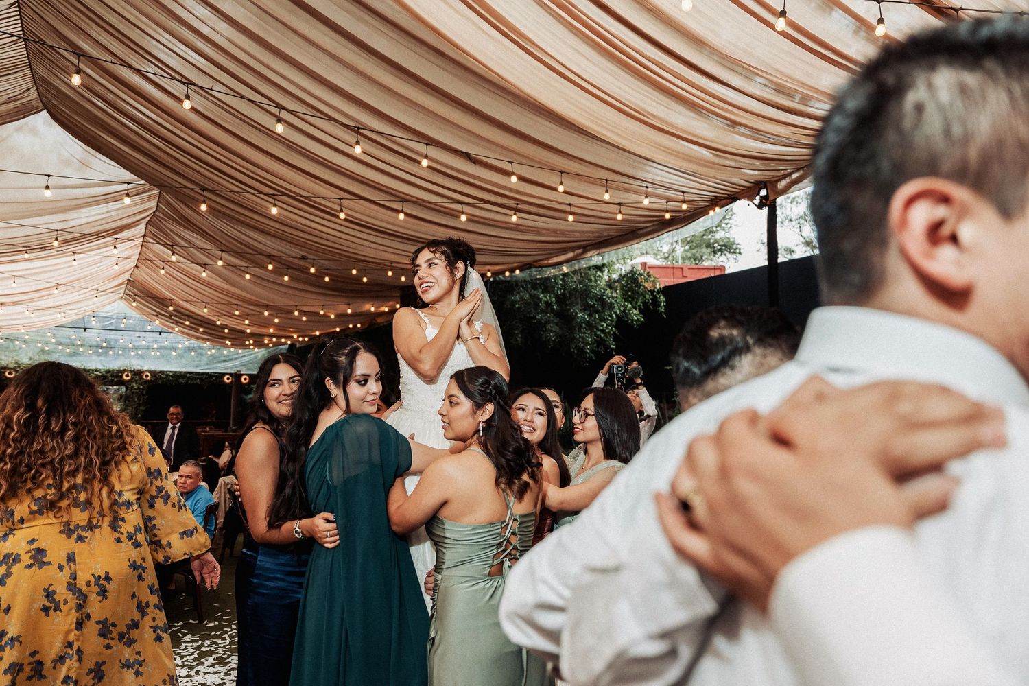 Guests celebrate at an outdoor wedding reception under draped fabric and string lights during daytime festivities.