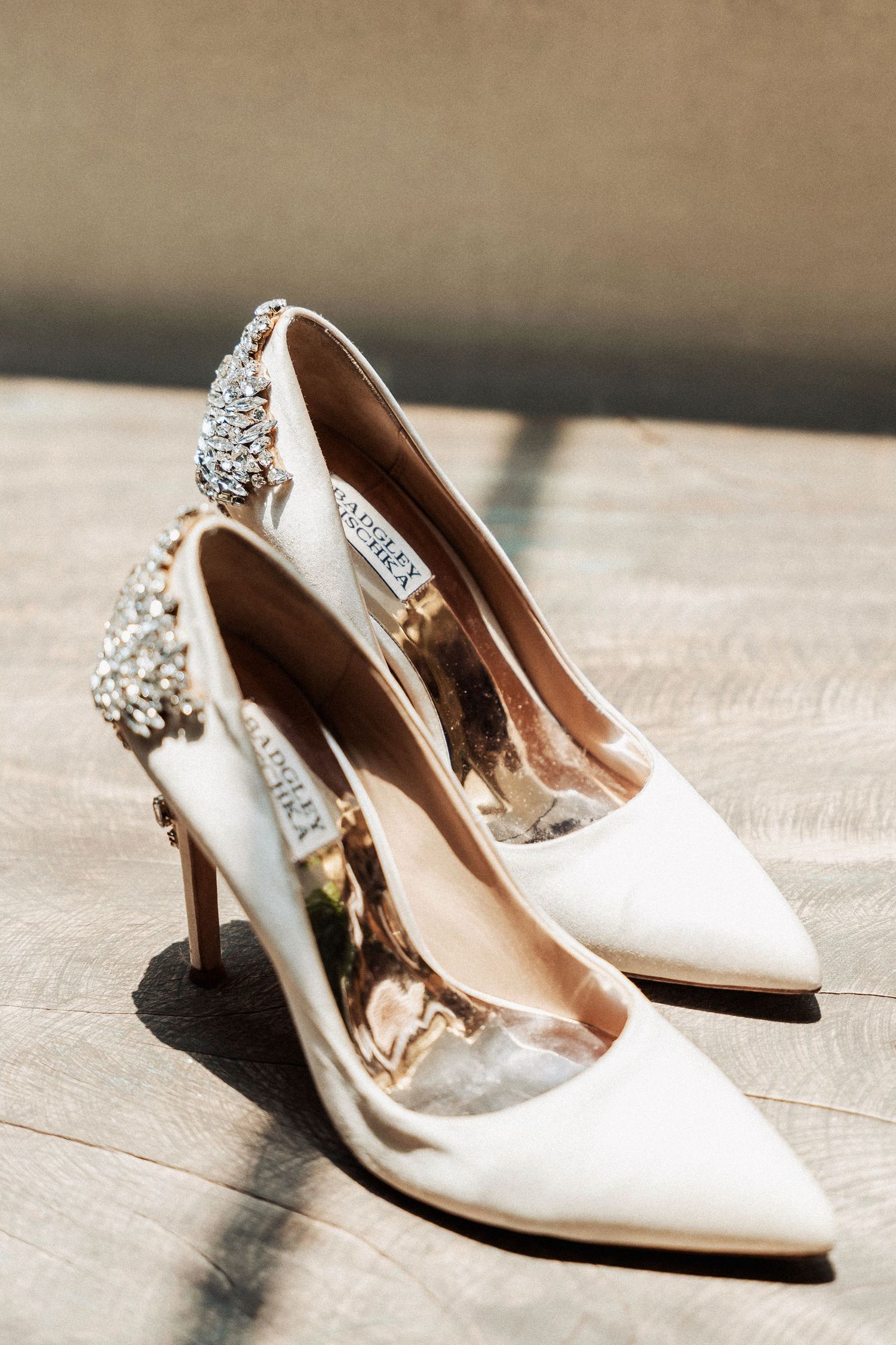 Elegant white satin bridal heels with crystal embellishments on the back displayed from different angles on wooden floor.
