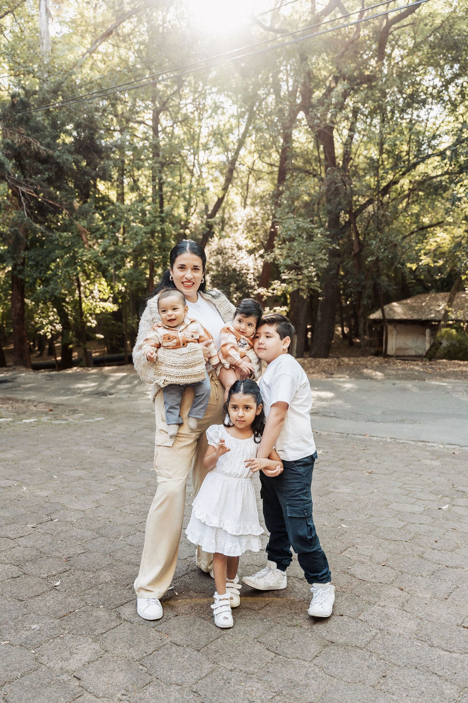 A family in coordinated white and neutral outfits poses together on a stone path with sunlit trees in the background.