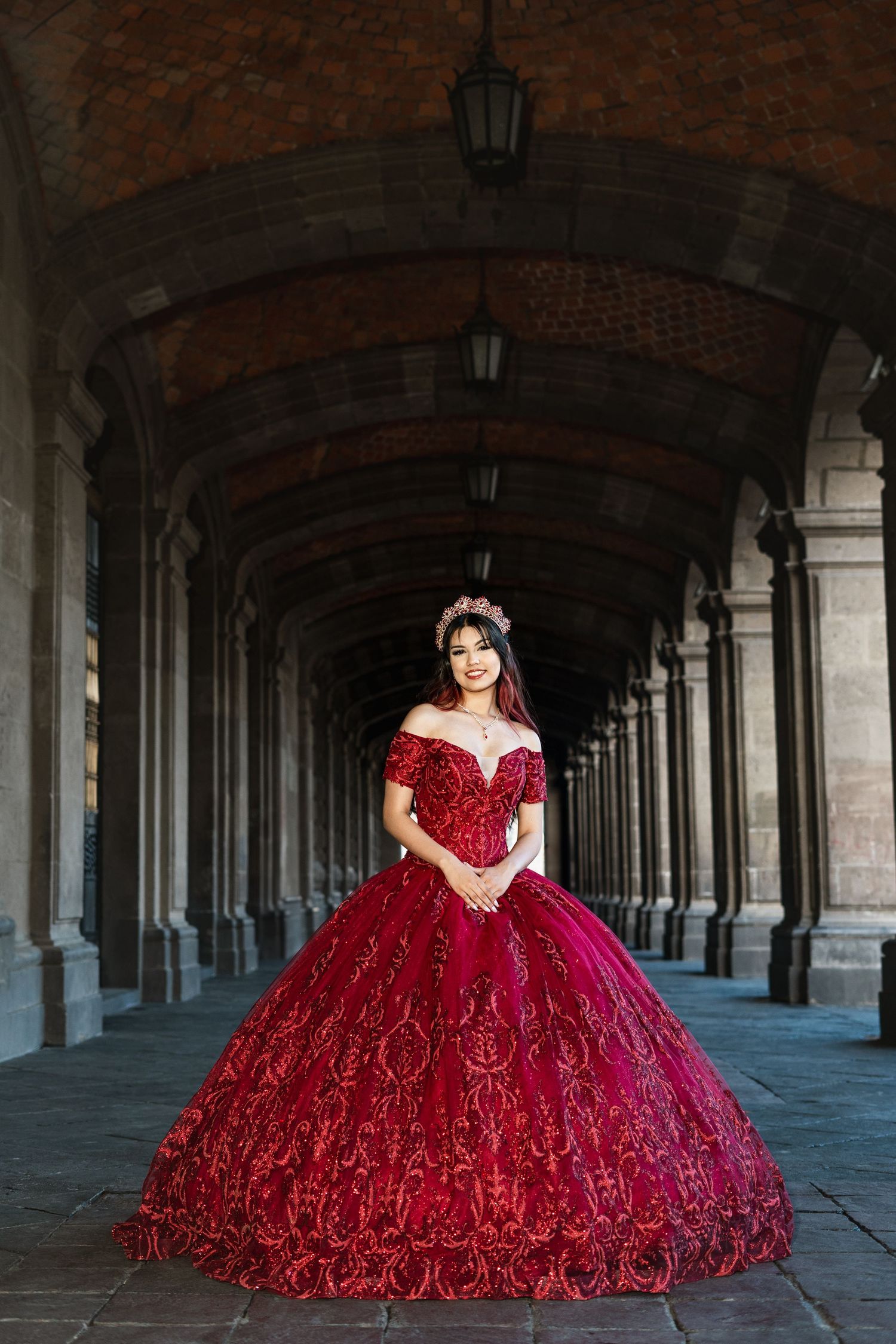 A stunning red ball gown with ornate details photographed under a dramatic arched corridor with hanging lanterns.