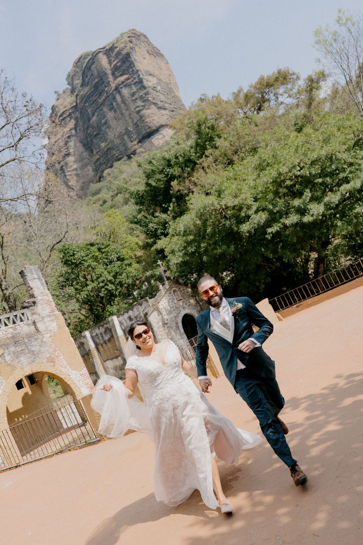 A couple in wedding attire running playfully on a path with a dramatic mountain peak rising in the background.