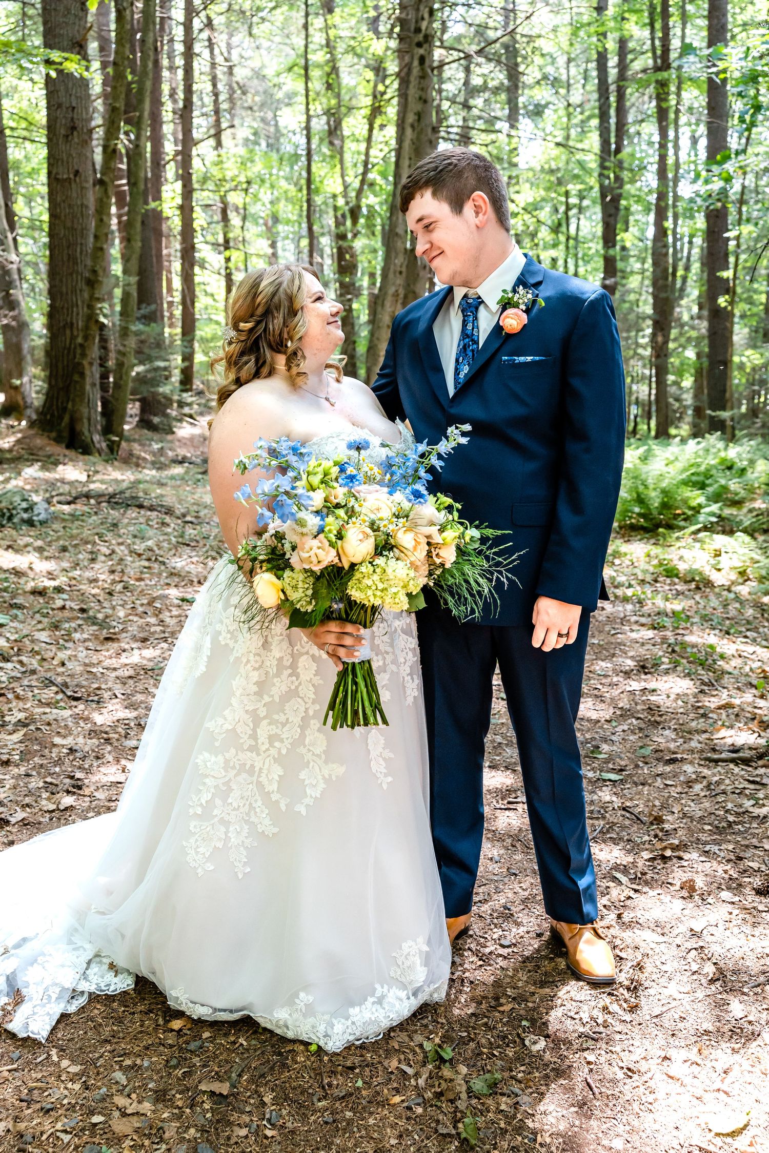 Wedding couple poses with yellow and blue bouquet against forest backdrop.