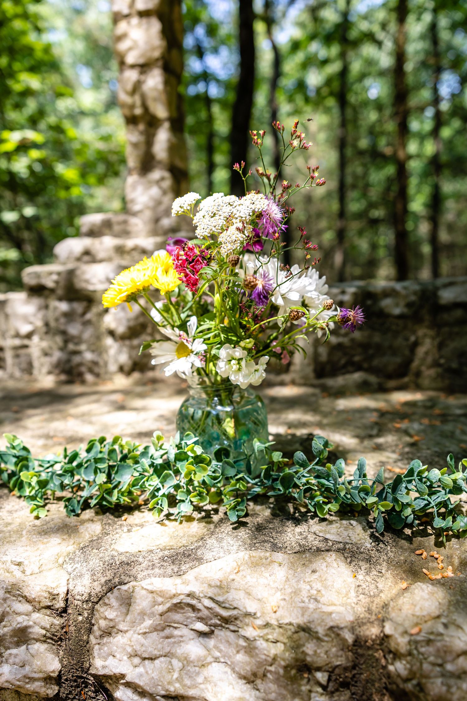 Colorful wildflower bouquet in mason jar placed on stone wall in forest setting.