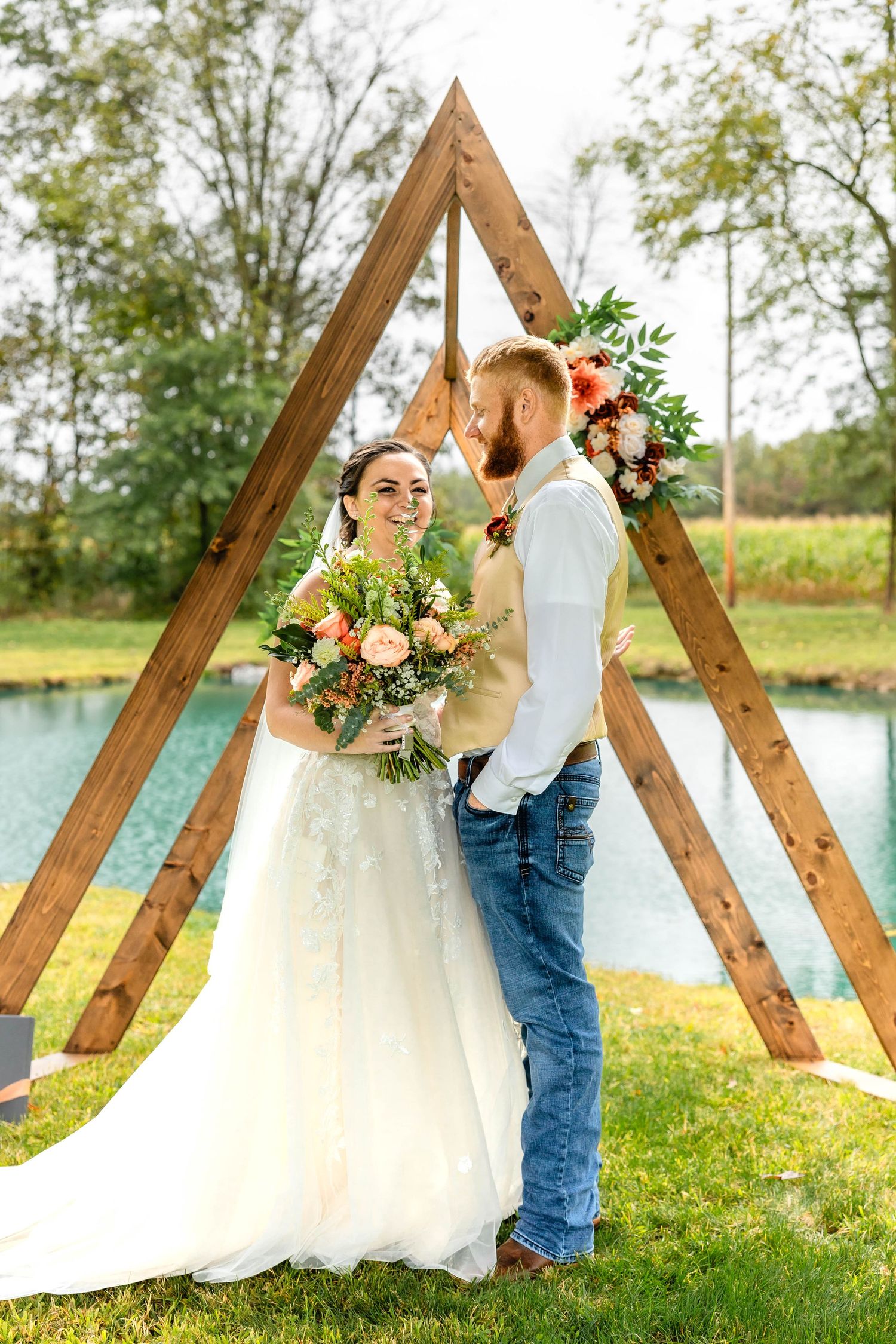 Newlyweds share a romantic moment under a wooden triangle arch decorated with flowers by a serene pond.