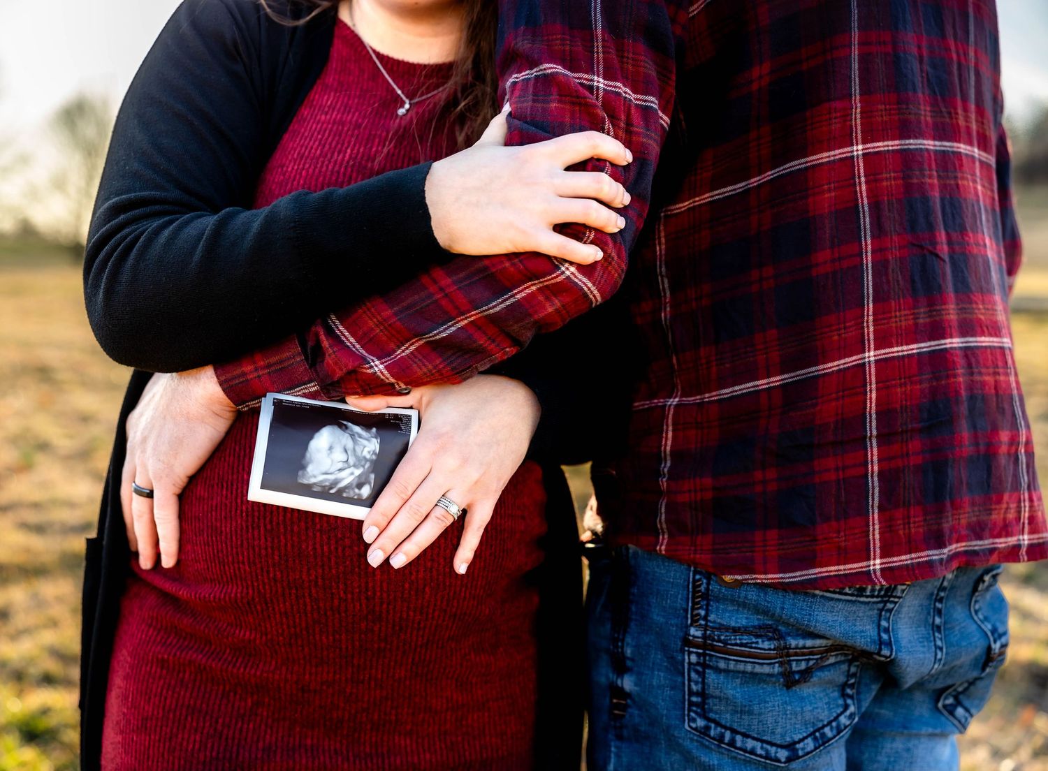 A couple holds a sonogram photo against a burgundy maternity dress in a field.
