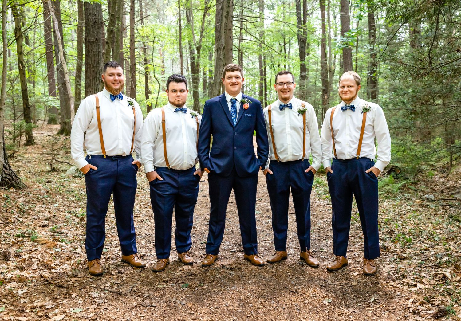 Groomsmen in navy pants and suspenders pose with groom in blue suit in a forest setting.