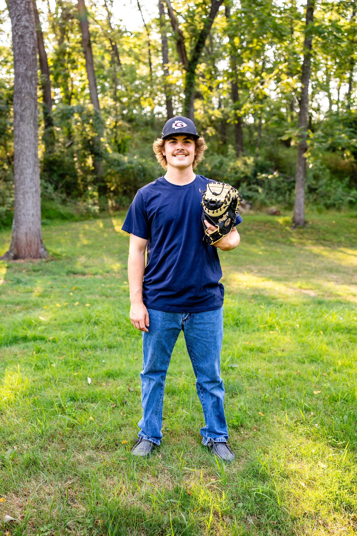 Baseball player in navy shirt and cap holds leather glove in wooded outdoor setting.