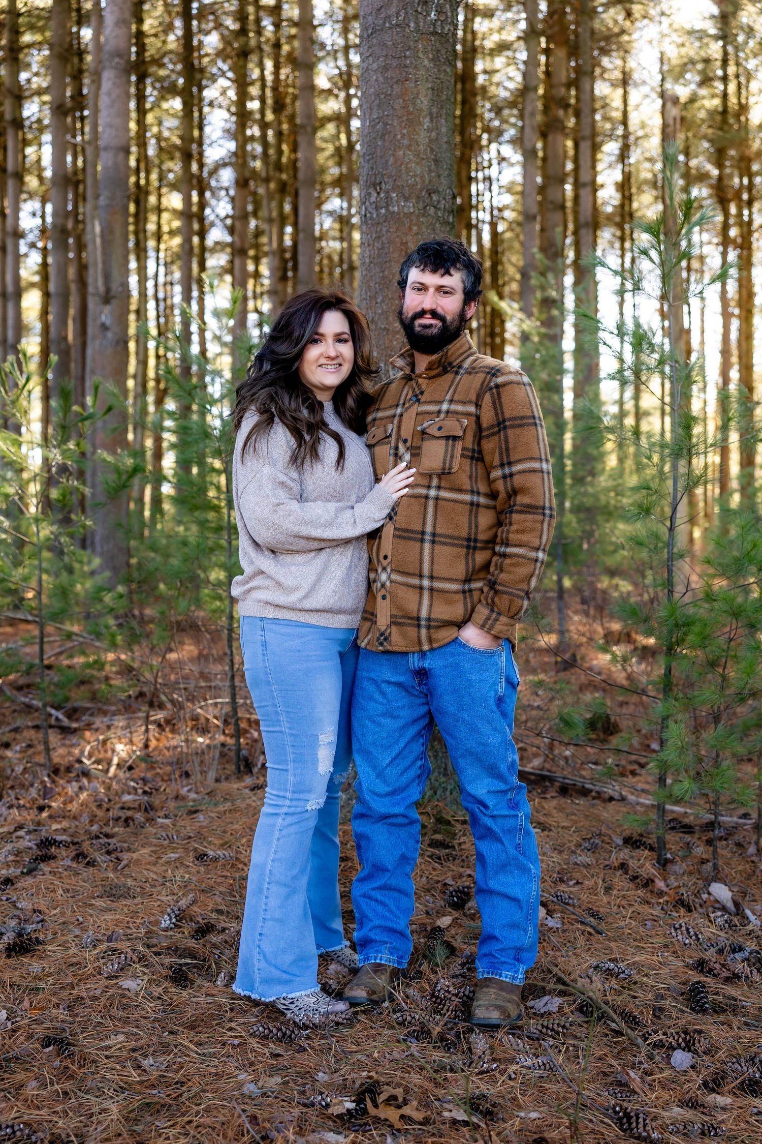 Couple standing together in casual attire among tall pine trees.