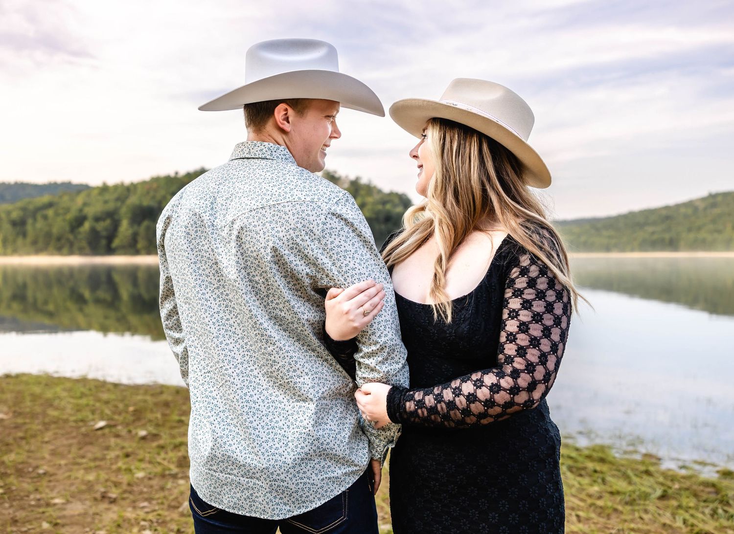 Couple in cowboy hats share intimate moment against mountain lake backdrop.
