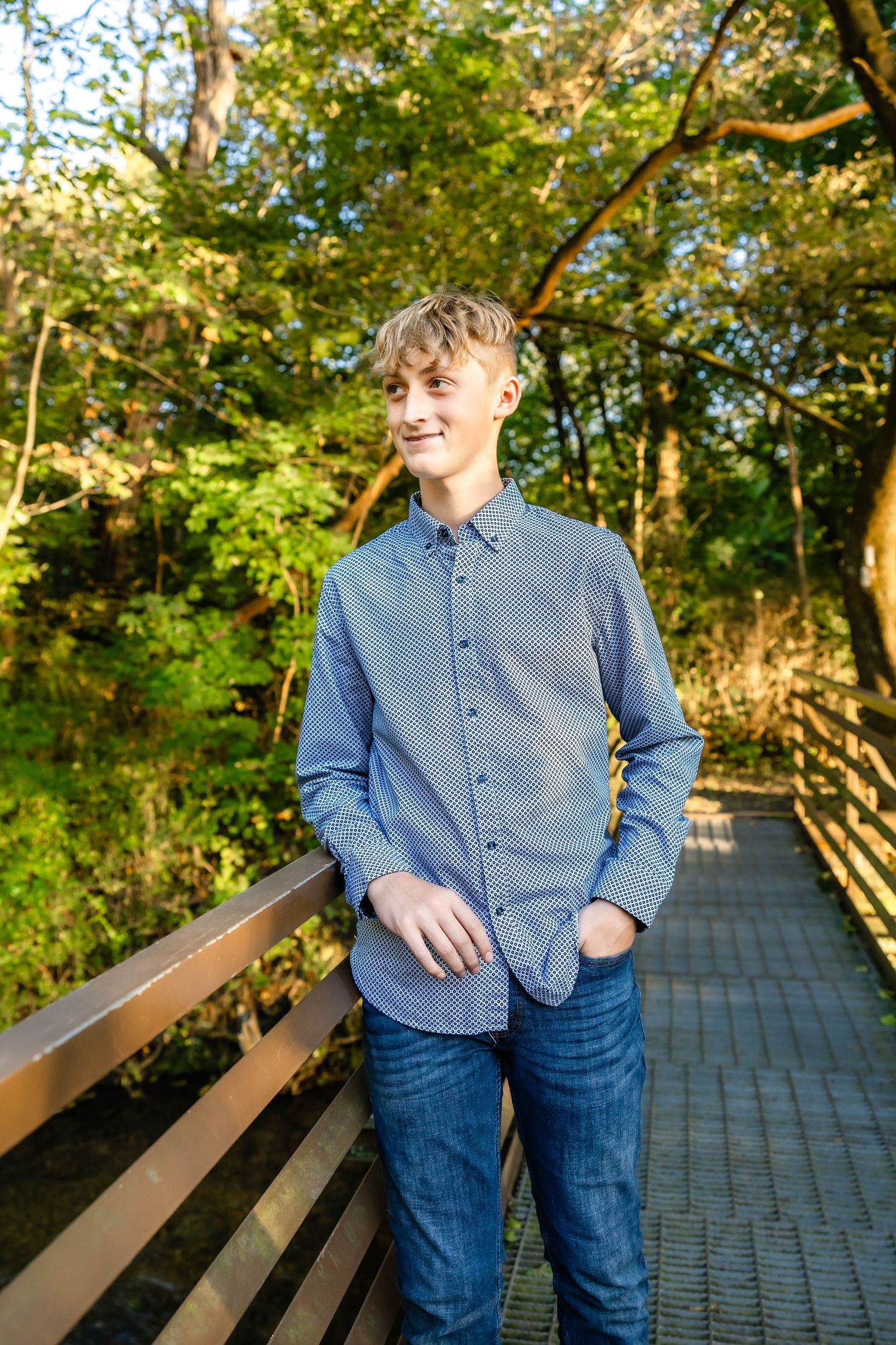 Person in casual blue button-down and jeans poses on wooden bridge with sunlit trees behind.
