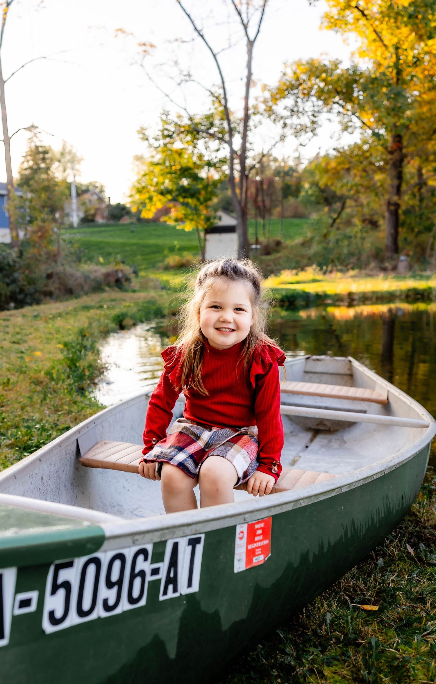 A child sits happily in a green canoe during autumn with fall foliage reflecting in the water.