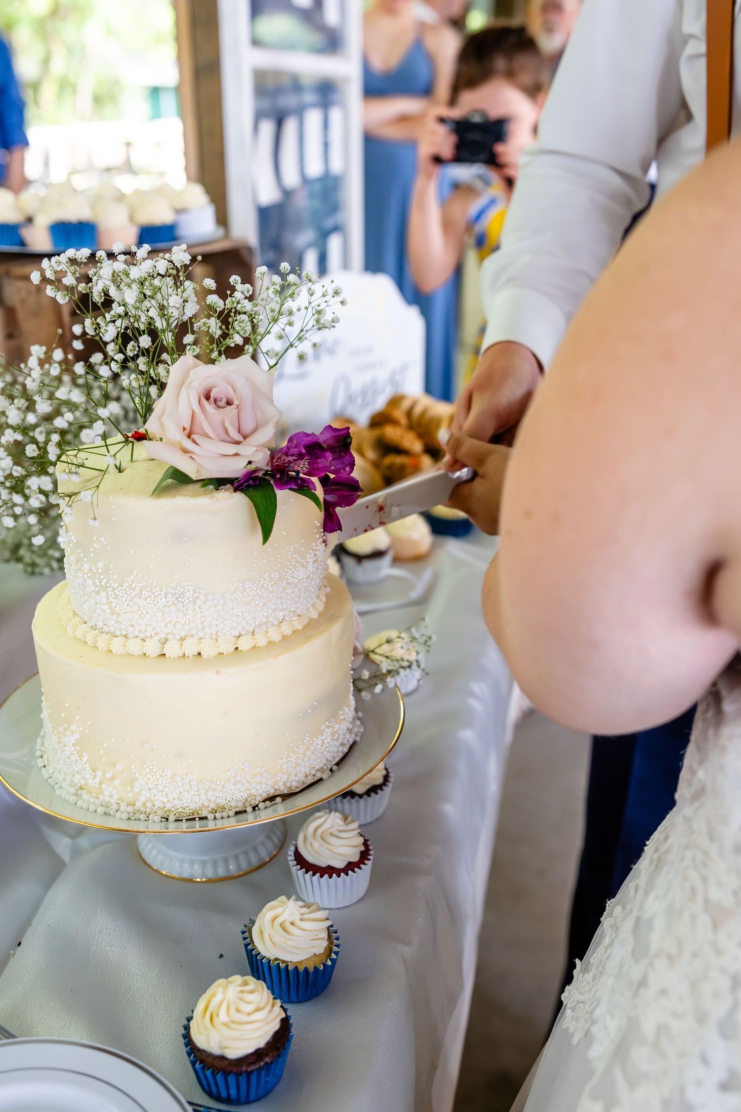 Wedding cake decorated with fresh flowers and pearls alongside cupcakes on a dessert table.