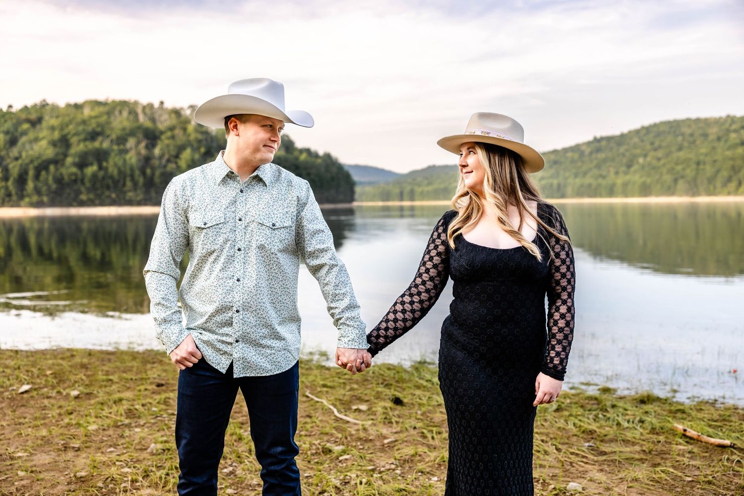 A couple in western attire holding hands by a scenic lake during their photo session.