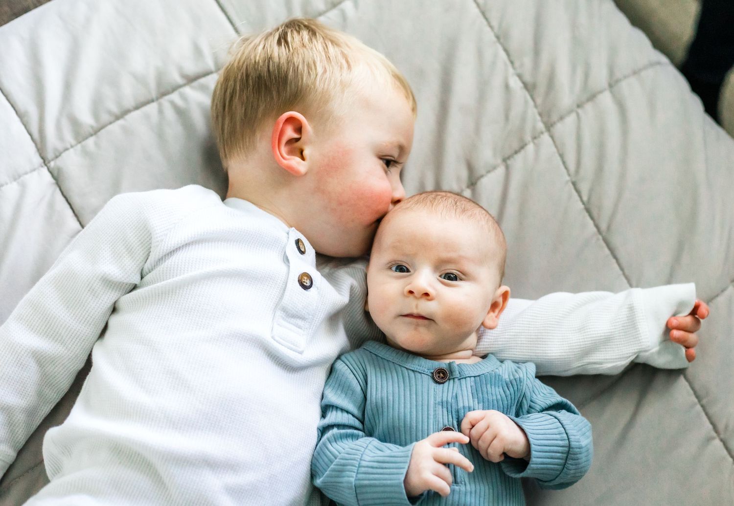 Sweet sibling moment with older child giving baby gentle kiss on quilted bedding.