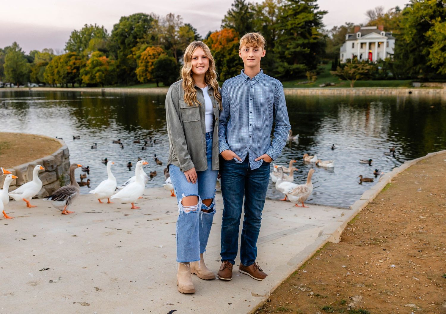 Couple standing near water with geese at lakeside during fall season.