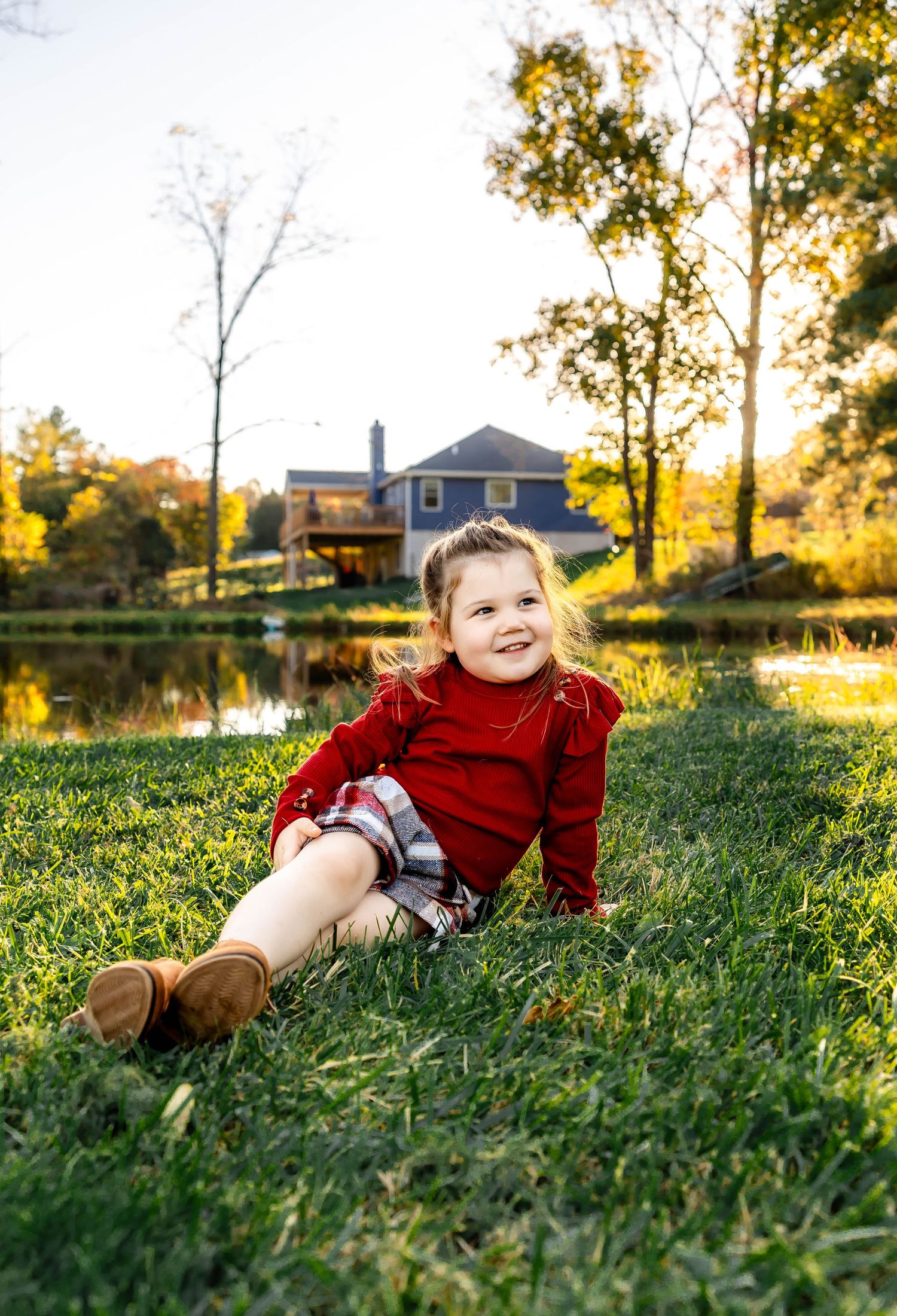 Child in red sweater reclines on grassy hill with blue house and pond in background during golden hour.