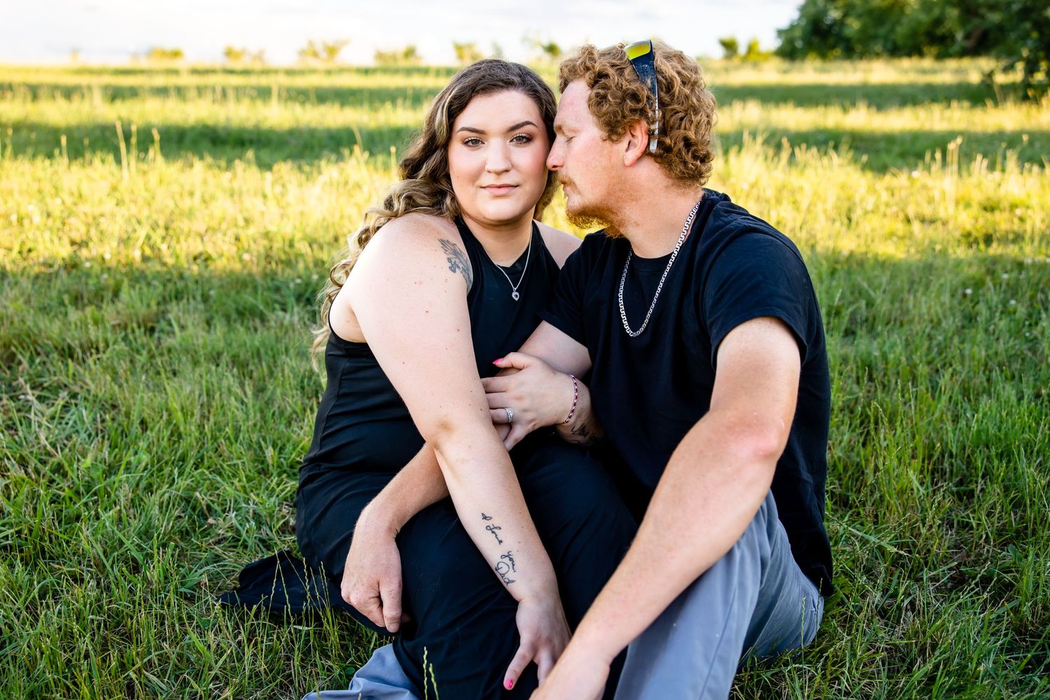 A couple in dark clothing embraces in a golden sun-lit field during sunset.