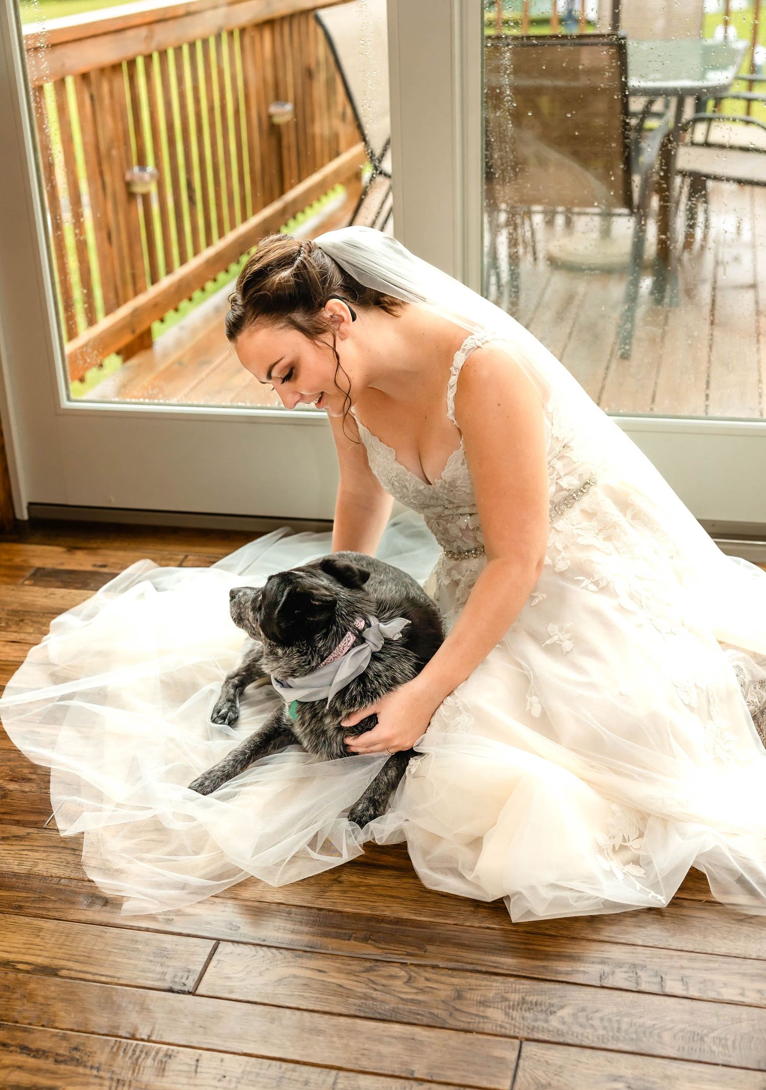 Bride in flowing white dress shares special moment with grey dog on hardwood floor.