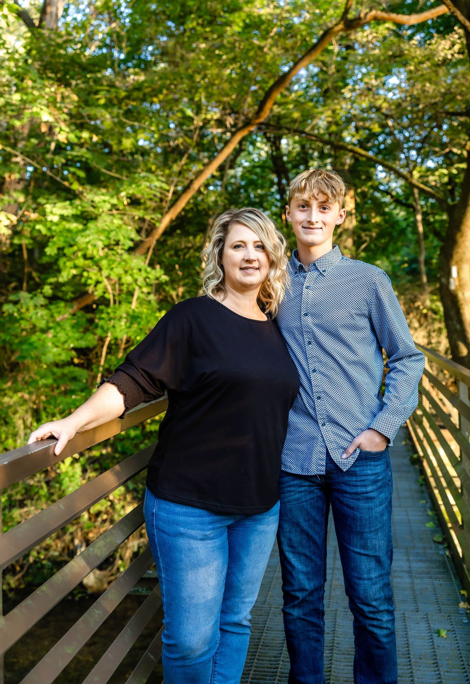 Two people in casual wear pose together on wooden bridge with autumn trees in background.