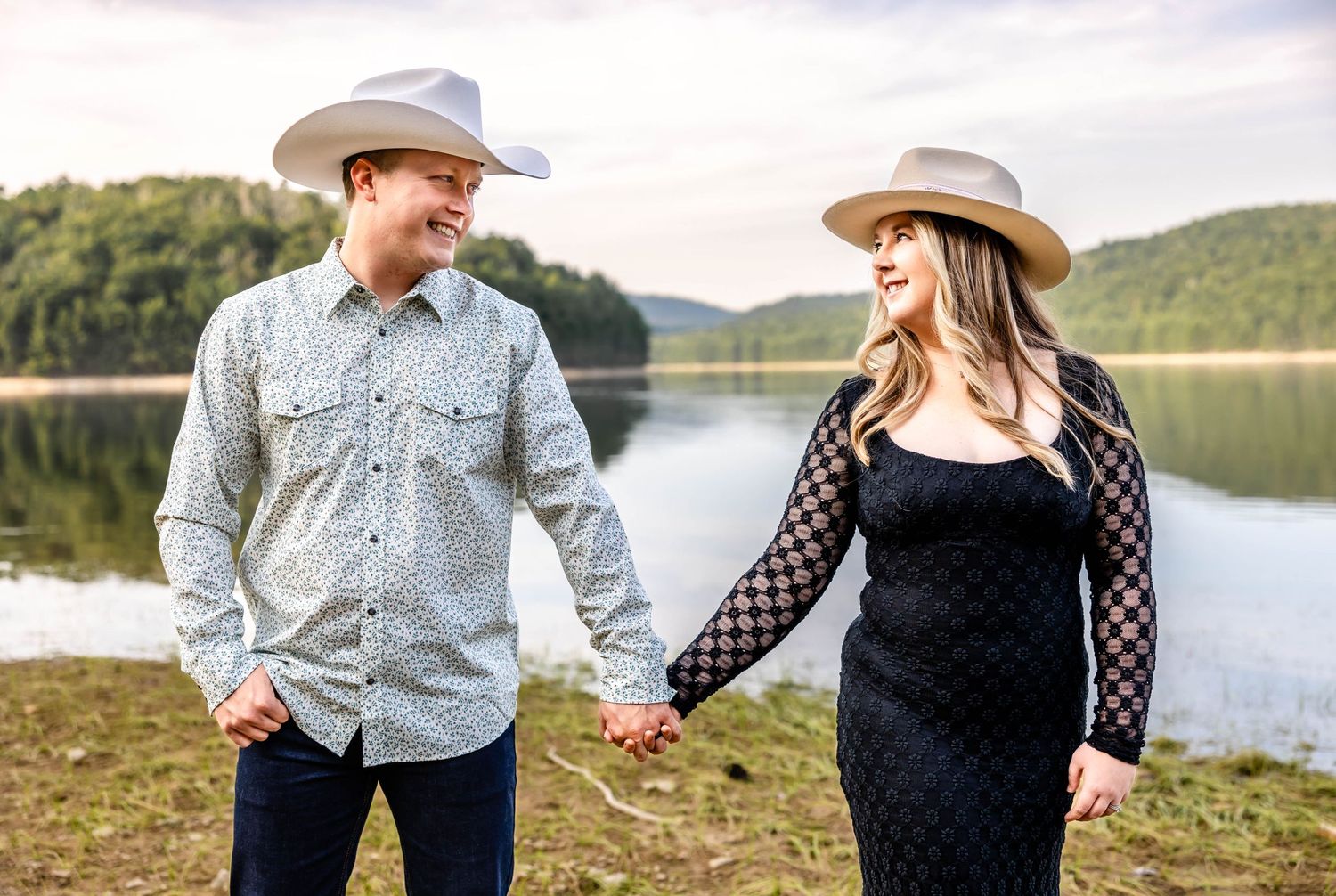 Couple gazes at each other while holding hands beside serene mountain lake.