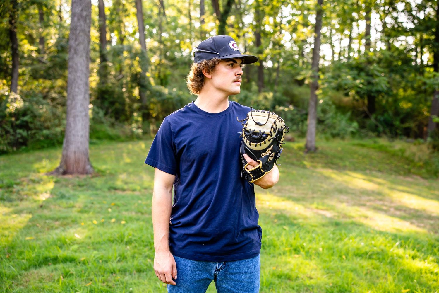 Baseball player poses with glove against wooded background in afternoon light.