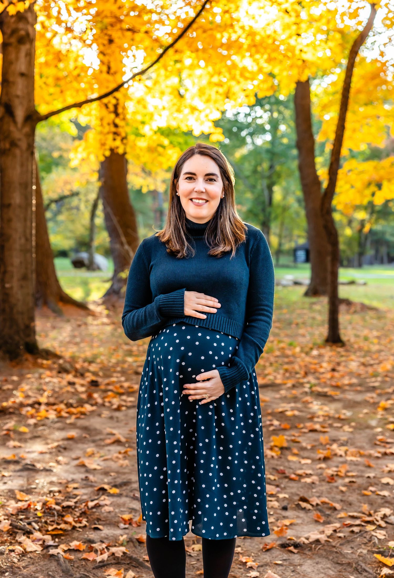 Maternity photo of woman in polka dot dress standing beneath golden fall trees.