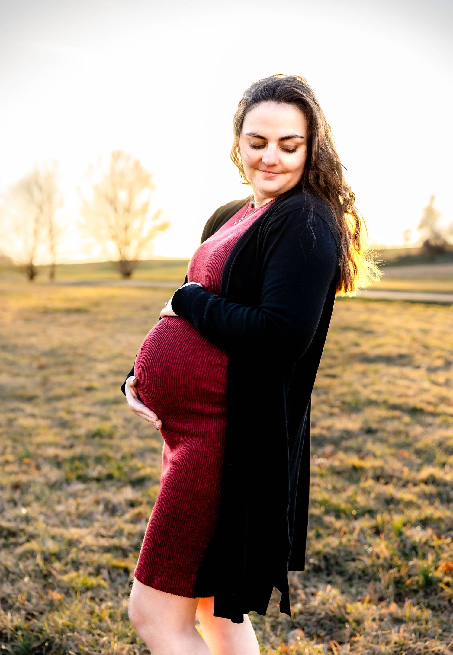 Expectant mother in burgundy dress and black cardigan smiling in golden sunlight.
