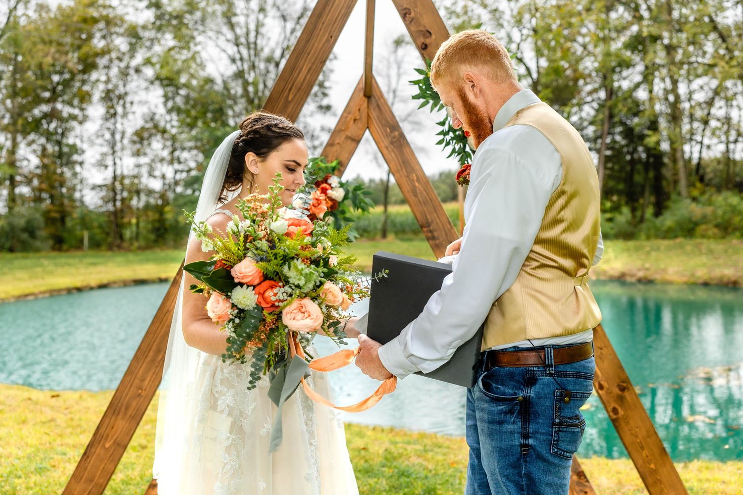 Wedding couple exchanges vows under a triangular wooden arch decorated with coral flowers beside a pond.
