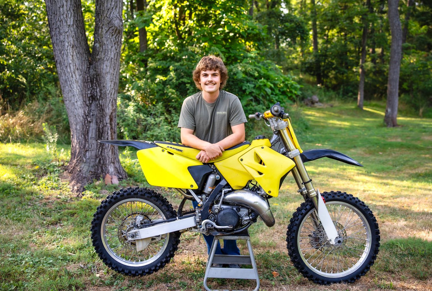 Yellow dirt bike displayed on stand with rider in gray shirt in wooded setting.