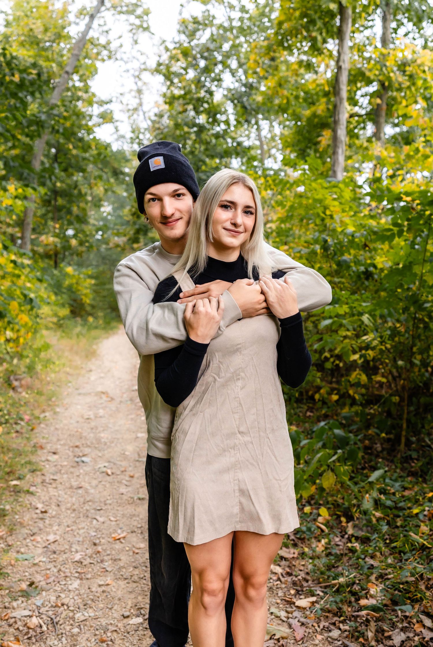 Couple embracing on forest path with fall foliage in background.
