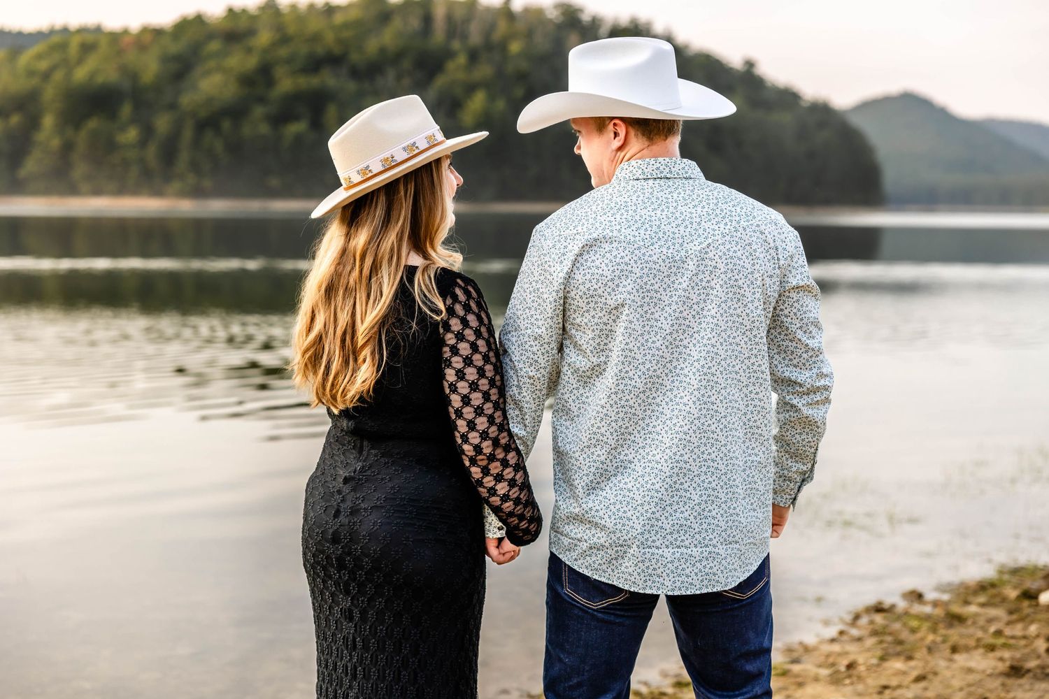 Romantic couple in western wear holding hands while standing by mountain lake.