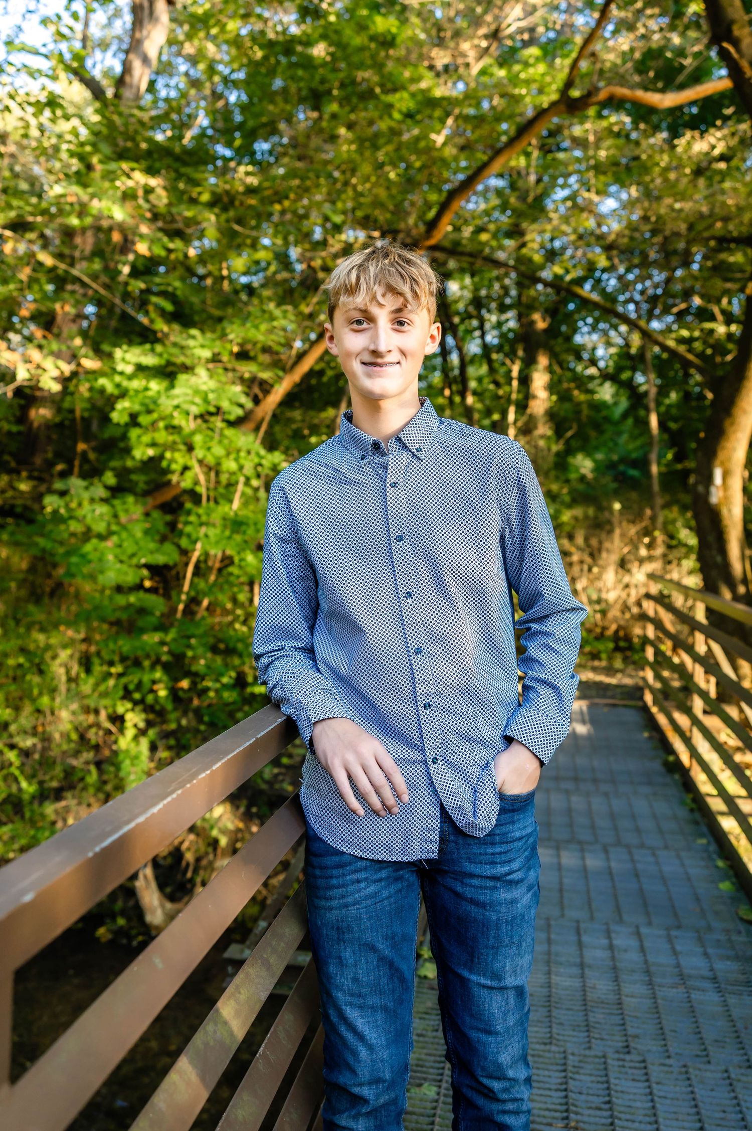 Young man in blue button-down shirt and jeans poses on wooden boardwalk with trees in background.