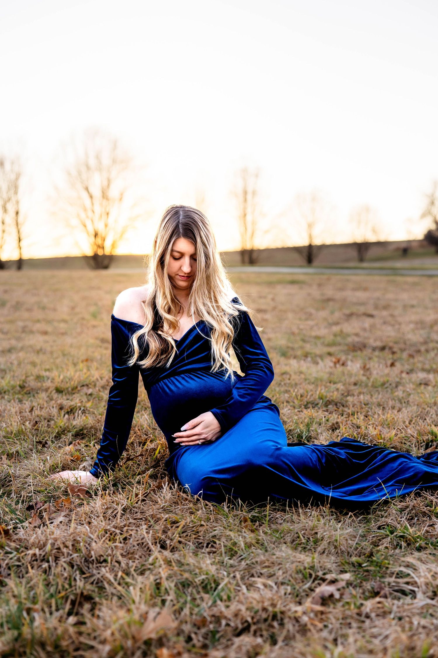 A person in a royal blue velvet maternity dress poses gracefully in a field at sunset.