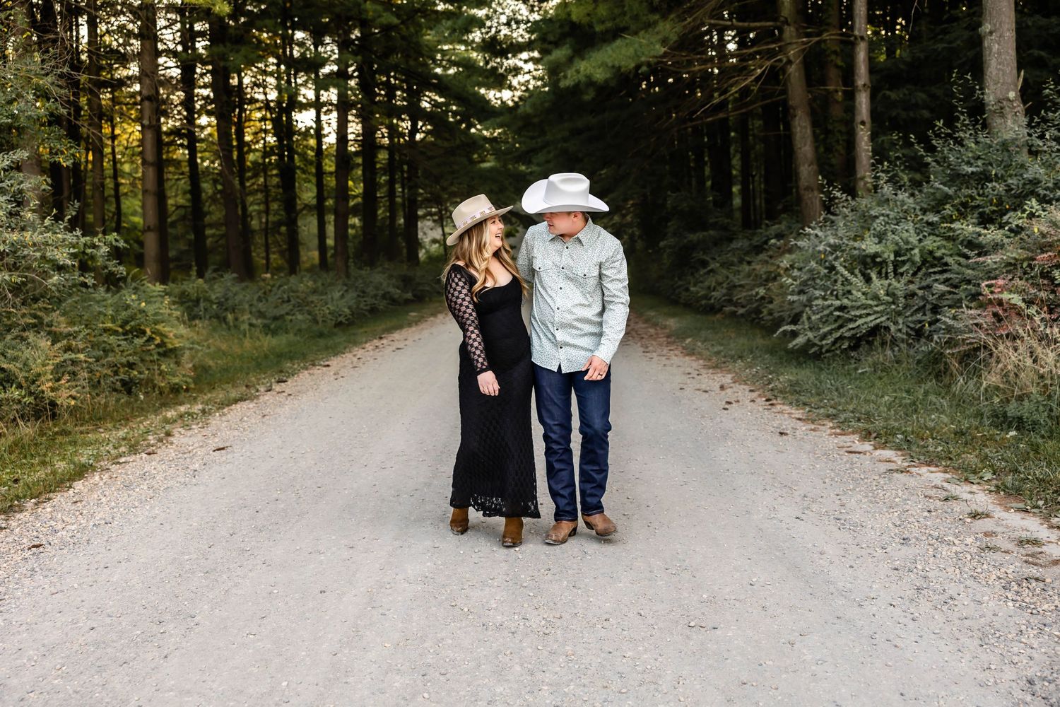A couple wearing cowboy hats stands close together on a forest trail.