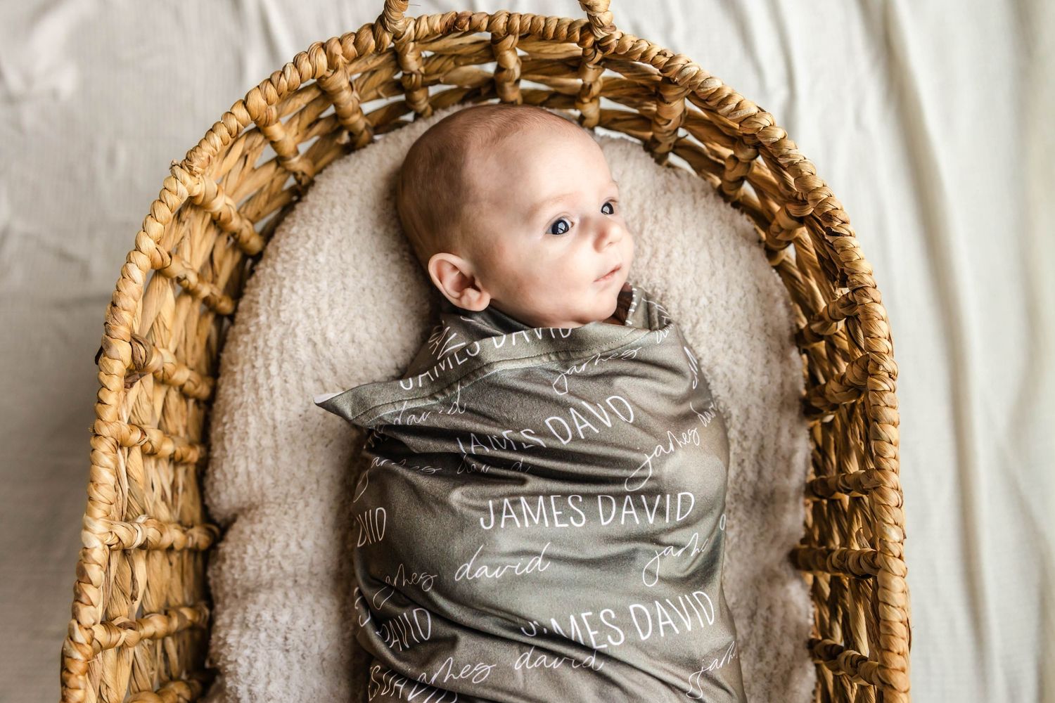 Infant wrapped in grey blanket resting in woven basket bassinet.